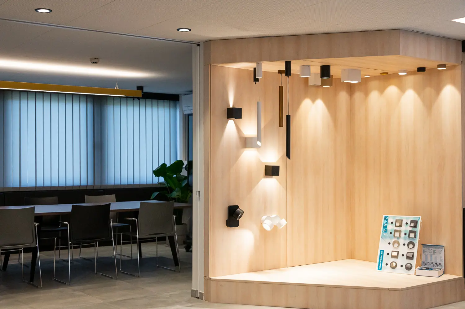 Vue combinée d'une salle de réunion de bureau derrière une vitre avec stores à gauche, et d'un présentoir d'éclairage dans une niche en bois allumée à droite