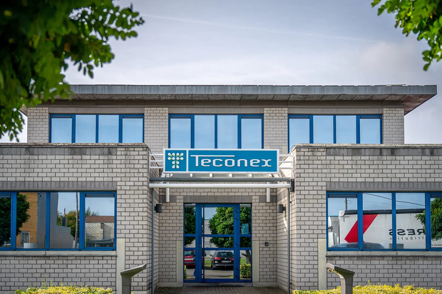 Façade extérieure complète du bâtiment de bureau principal de Teconex, un bâtiment en briques gris clair à deux étages avec des fenêtres bleues et le logo "Teconex" bleu au-dessus de l'entrée